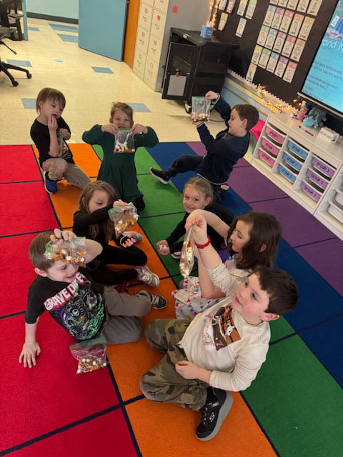 students seated on classroom rug holding up crafts