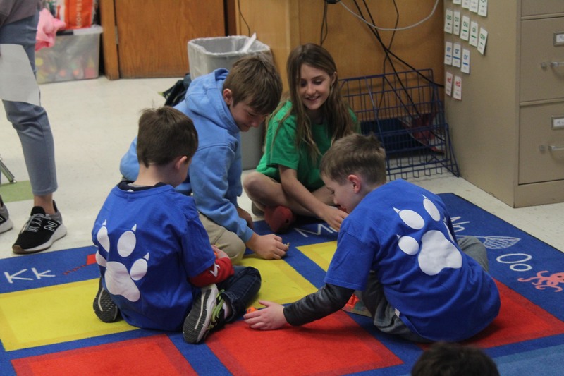 three boys and girl sitting on classroom rug playing game