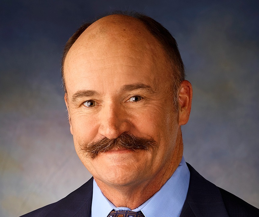 A mans picture from the waist up wearing a suit and tie. He also has a mustache on his face.