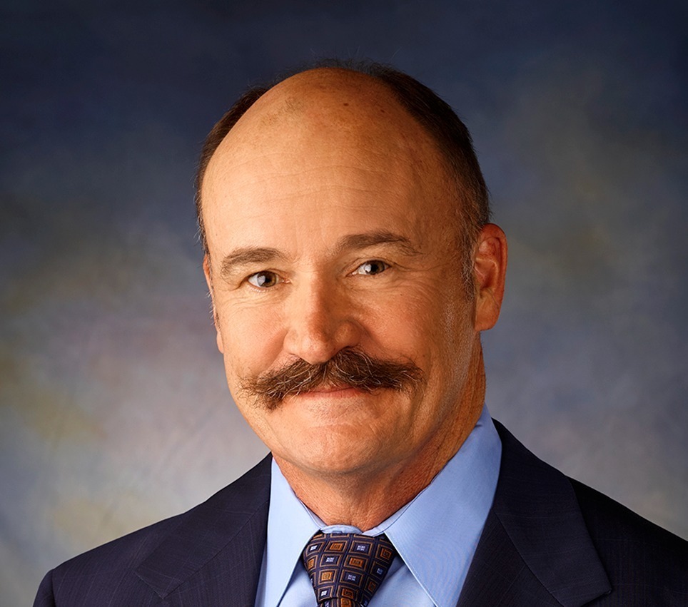 A mans picture from the waist up wearing a suit and tie. He also has a mustache on his face.