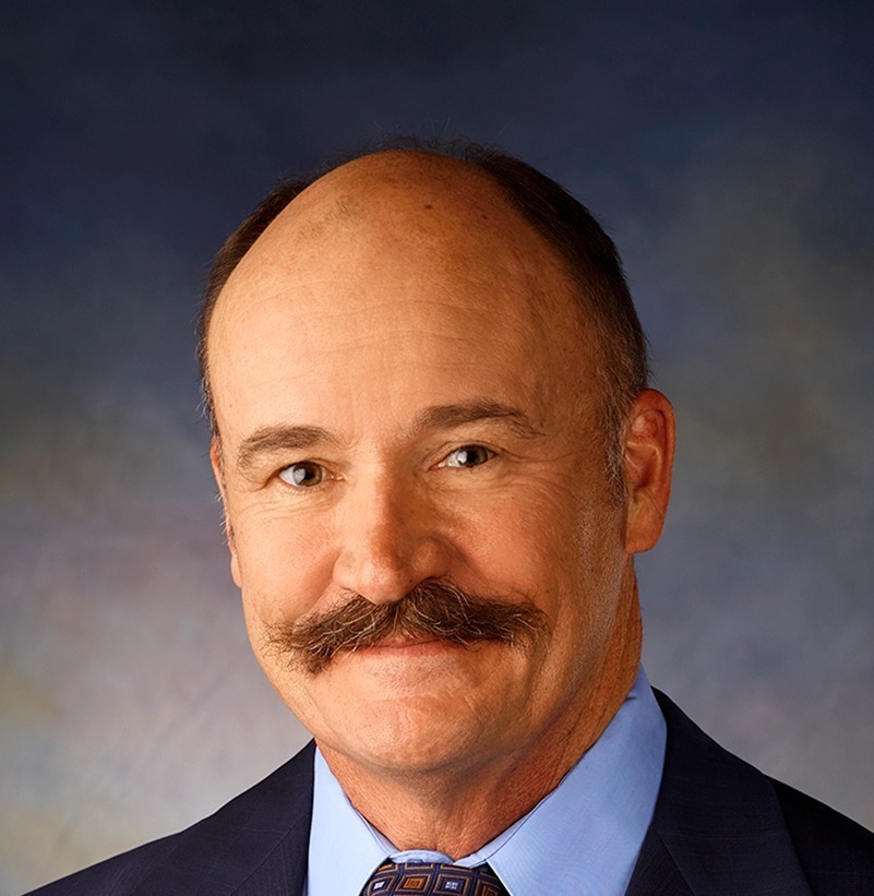 A mans picture from the waist up wearing a suit and tie. He also has a mustache on his face.