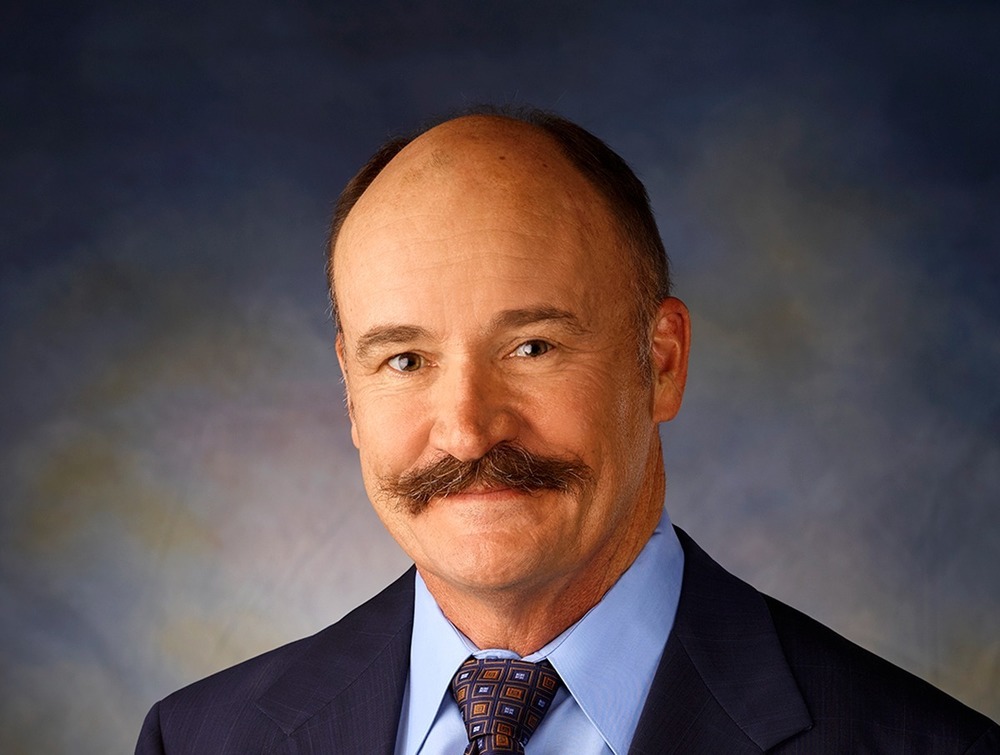 A mans picture from the waist up wearing a suit and tie. He also has a mustache on his face.