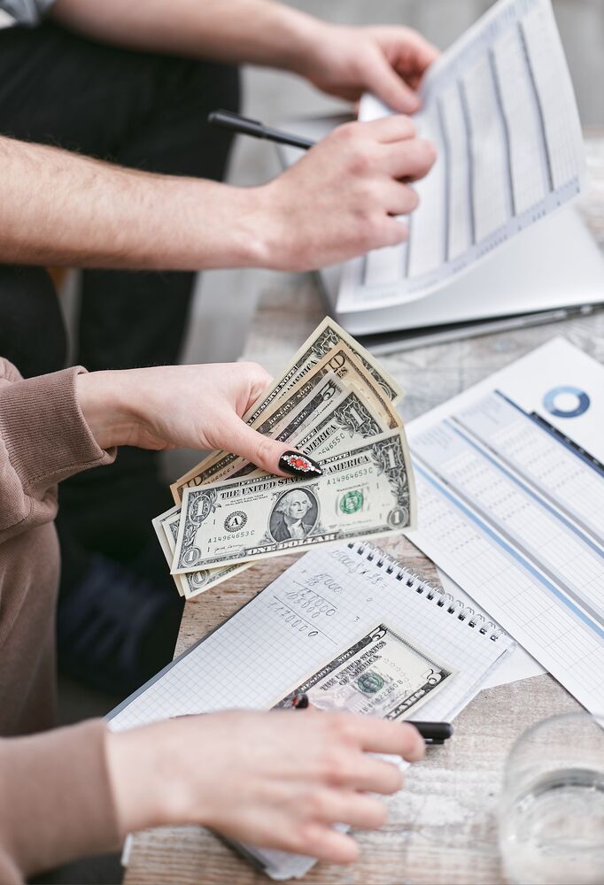 Spreadsheets and cash money laid out on a table and two peoples hands are holding them and it looks like they are working with them