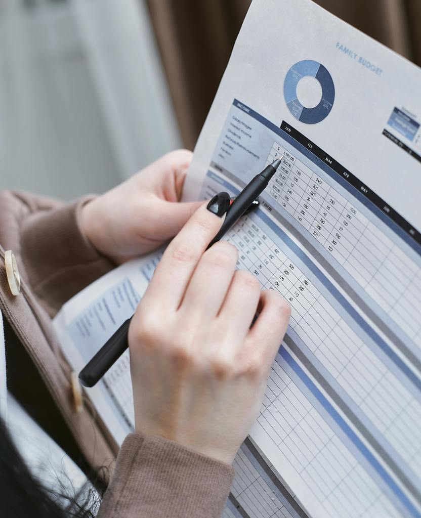 someone pointing to a spreadsheet with a pen in their hand. The hand is light colored with black nail polish. 