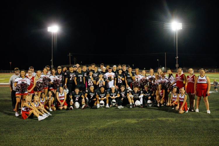 photo on a football at night. pictured is the high school football team, coaches, 5/6, JH & HS Cheer. Aaron Hoggard is in the middle holding a plaque honoring 20 years of dedication to the Chiefs