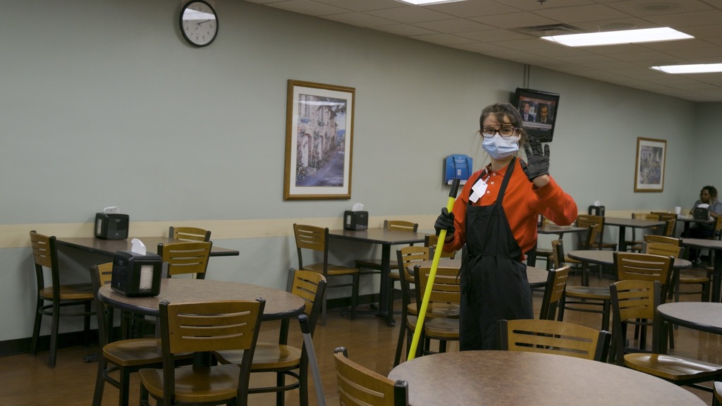 girl with broom in cafeteria waving at camera