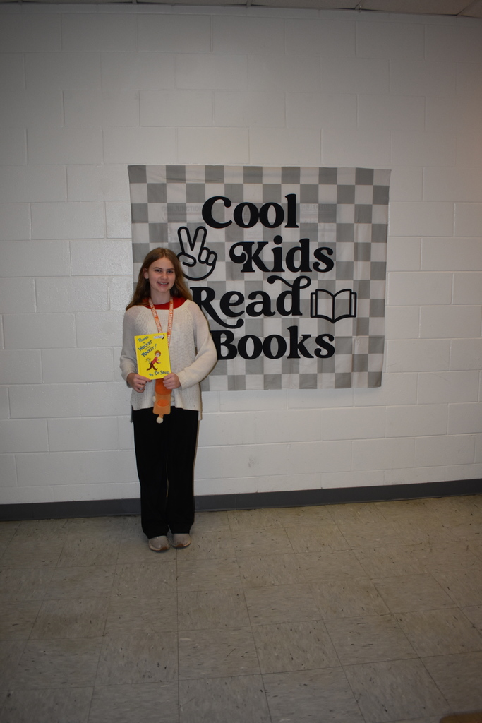 A student standing with her favorite Dr. Seuss book in front of a sign that says "cool kids Read books:"