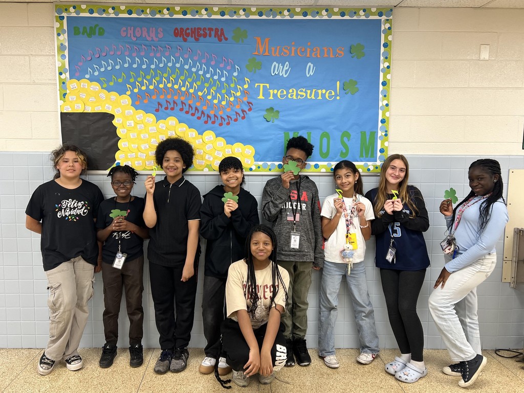 Group of students holding paper clovers and standing in front of thier bulletin oard design of music notes an text that says musicians are a treasure