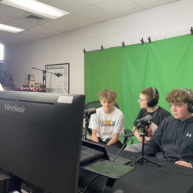 Three middle school students sit at a table in front of a green screen in a media center, wearing headphones and speaking into microphones while recording a podcast. Studio lights and a computer monitor are set up in front of them.
