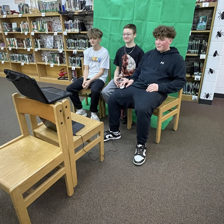 Three middle school students sit at a table in front of a green screen in a media center, wearing headphones and speaking into microphones while recording a podcast. Studio lights and a computer monitor are set up in front of them.