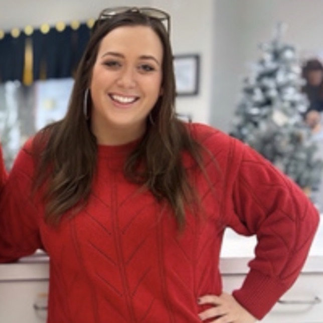 A staff member stands in a school front office wearing a red sweater and black pants, smiling in a welcoming environment.