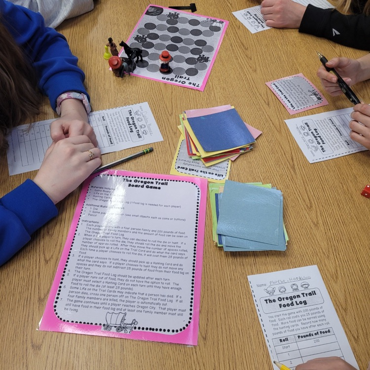 Students sit at desks in a classroom playing a paper-based version of the Oregon Trail game, using worksheets and materials to make decisions and track their journey while learning about westward expansion.