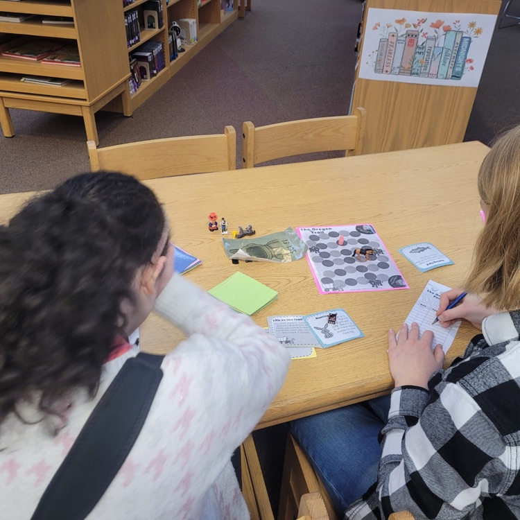 Students sit at desks in a classroom playing a paper-based version of the Oregon Trail game, using worksheets and materials to make decisions and track their journey while learning about westward expansion.