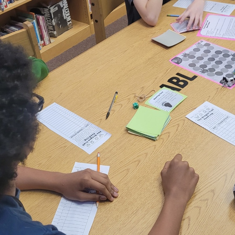 Students sit at desks in a classroom playing a paper-based version of the Oregon Trail game, using worksheets and materials to make decisions and track their journey while learning about westward expansion.