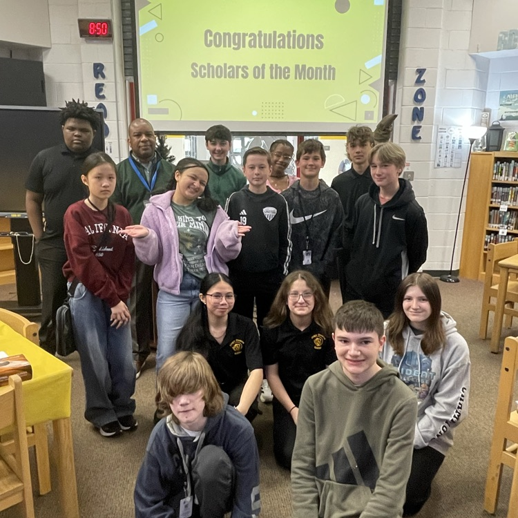 Students celebrating at a Scholar of the Month breakfast, posing with donuts in front of a commemorative banner.