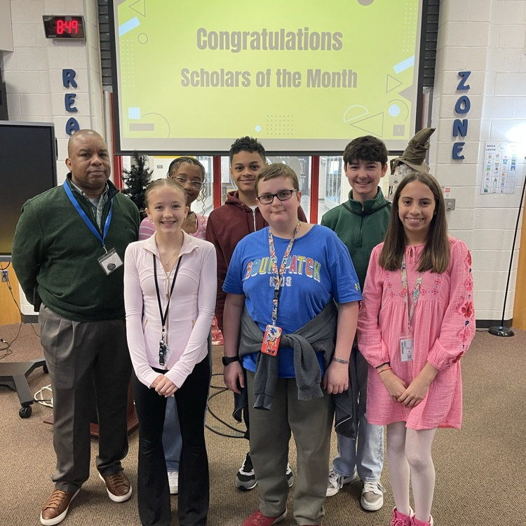 Students celebrating at a Scholar of the Month breakfast, posing with donuts in front of a commemorative banner.
