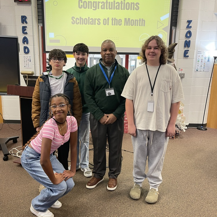 Students celebrating at a Scholar of the Month breakfast, posing with donuts in front of a commemorative banner.