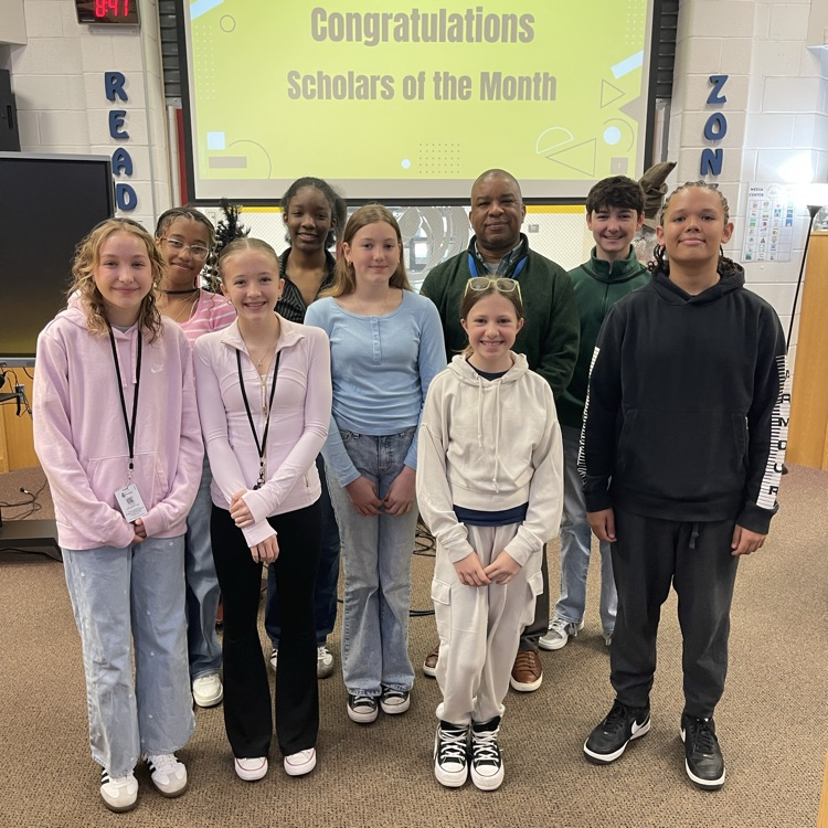 Students celebrating at a Scholar of the Month breakfast, posing with donuts in front of a commemorative banner.