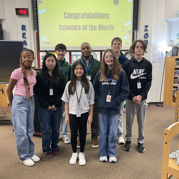 Students celebrating at a Scholar of the Month breakfast, posing with donuts in front of a commemorative banner.