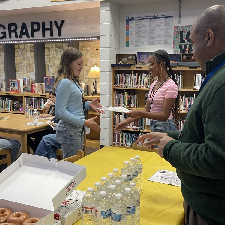 Students celebrating at a Scholar of the Month breakfast, posing with donuts in front of a commemorative banner.