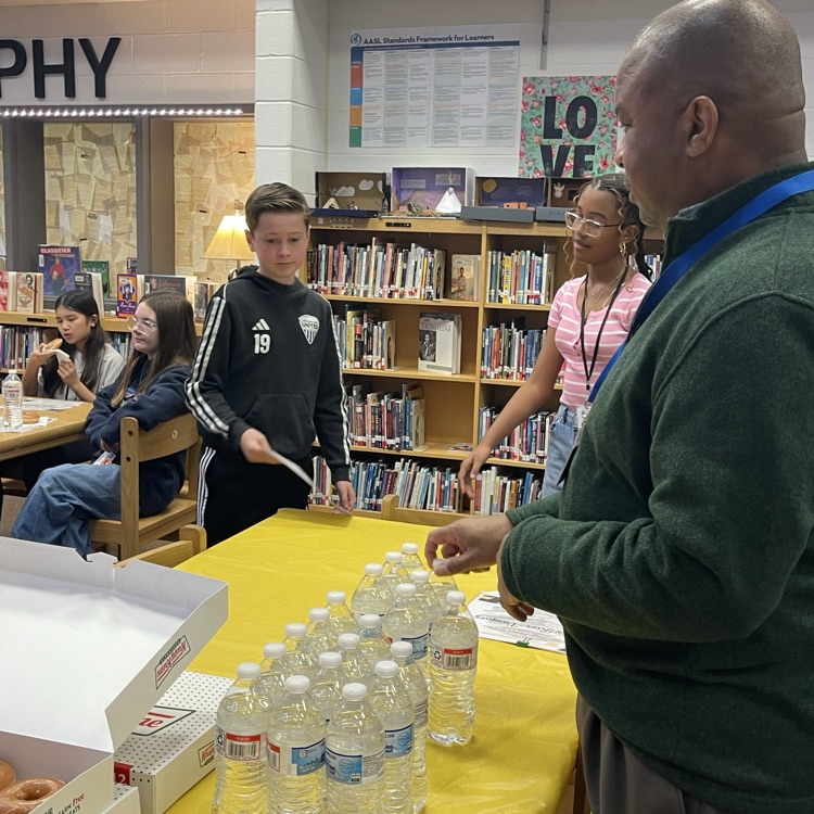 Students celebrating at a Scholar of the Month breakfast, posing with donuts in front of a commemorative banner.