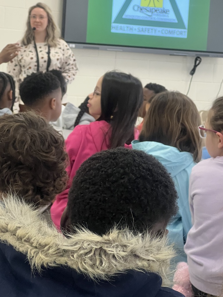 A presenter speaks to a group of elementary students seated closely together, while a screen behind her displays a Chesapeake Mosquito Control presentation.
