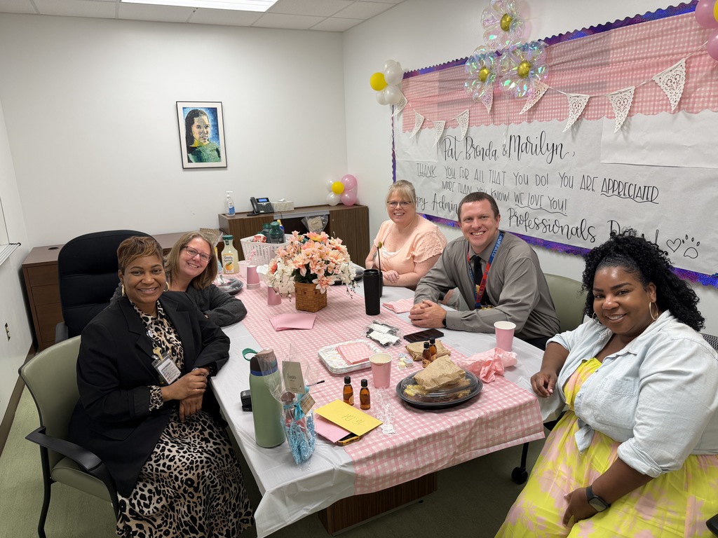 Five staff members sit around a small table in a decorated office, smiling at the camera during an administrative professionals appreciation celebration. The table is covered with a pink gingham tablecloth and holds flowers, snacks, drinks, and small gifts. Behind them, a large handmade banner with pink decorations and balloons thanks our administrative professionals for all they do.