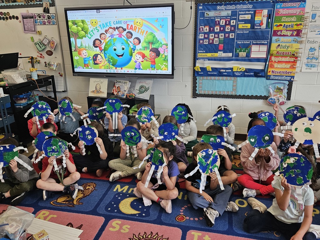 A group of kindergarteners sitting on a colorful classroom rug, holding handmade paper Earth crafts with googly eyes and folded paper arms over their faces. A screen in the background reads "Let's take care of Earth!".