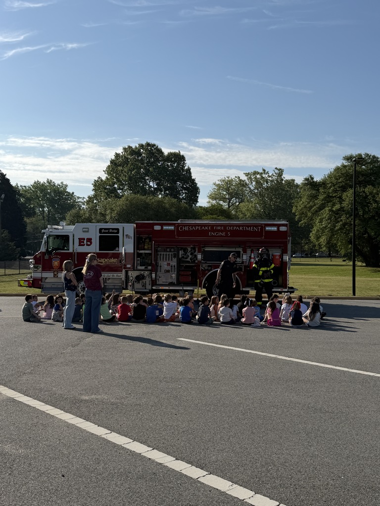 Class of students seated outside, listening attentively to firefighters speaking in front of a fire truck during Career Day