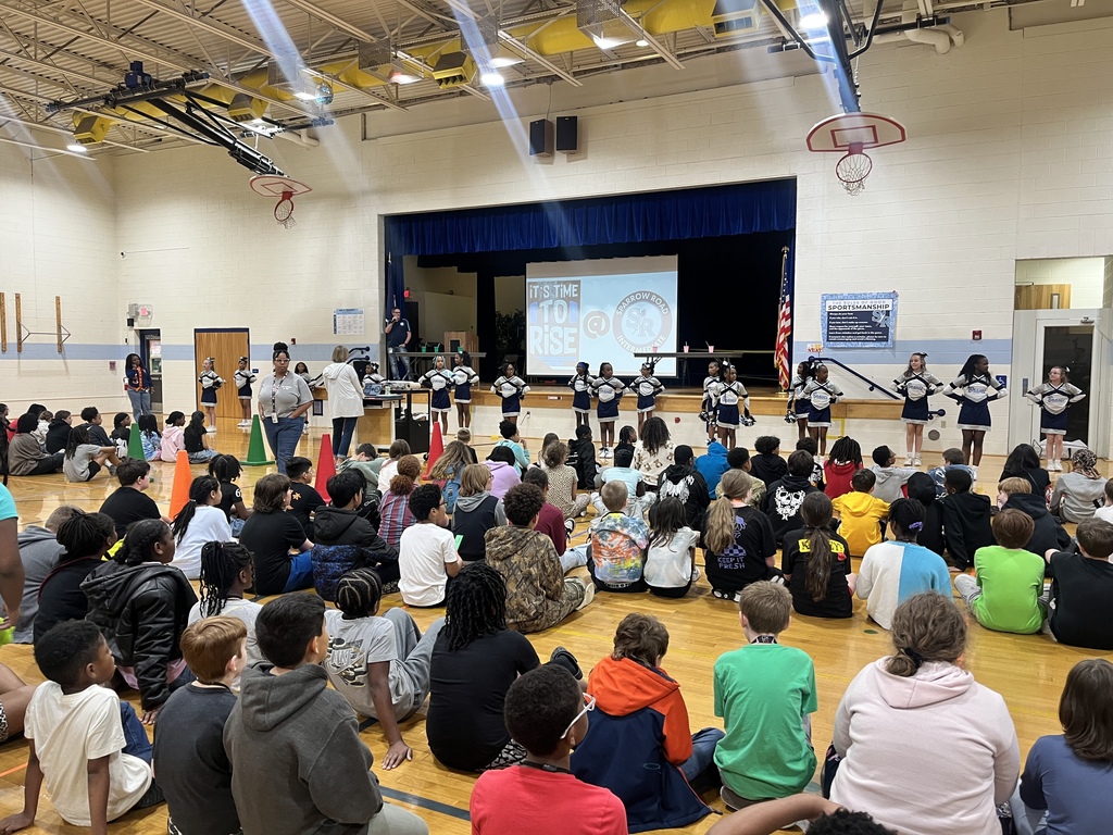 Students all sitting in the gym while Admin presents on stage with cheerleaders upfront.