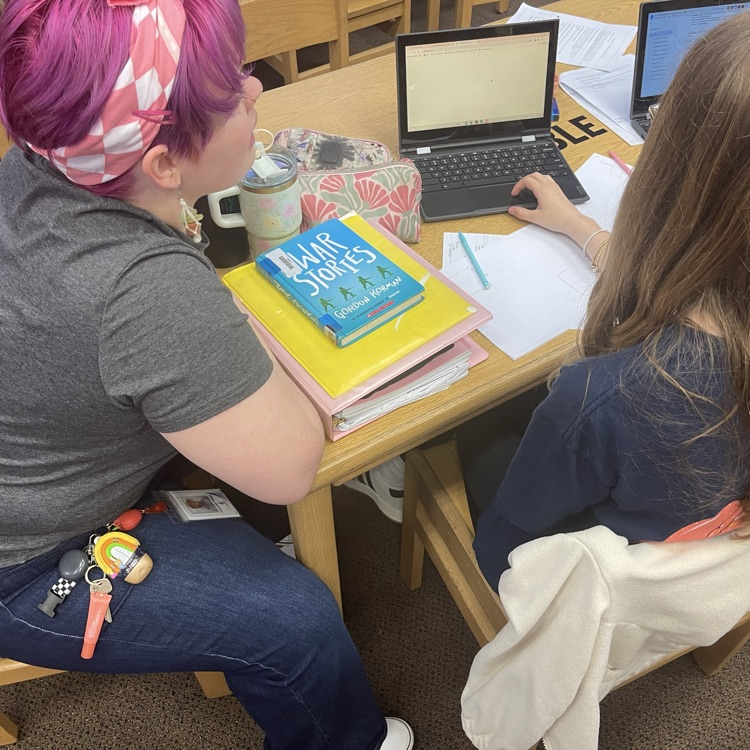 high-angle shot of two students working collaboratively at a library table. They are surrounded by printed research papers and are using laptops to type up their findings.
