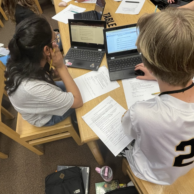 high-angle shot of two students working collaboratively at a library table. They are surrounded by printed research papers and are using laptops to type up their findings.