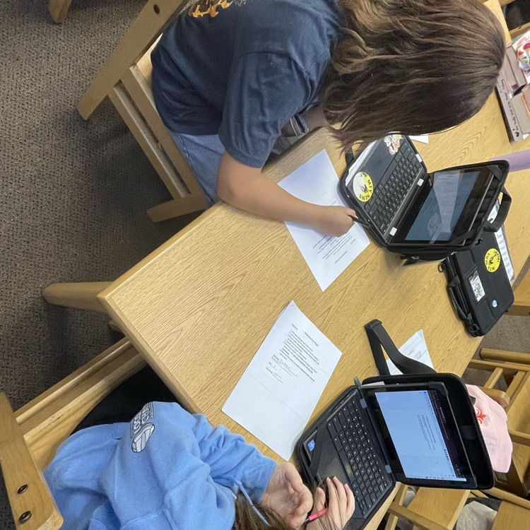 high-angle shot of two students working collaboratively at a library table. They are surrounded by printed research papers and are using laptops to type up their findings.