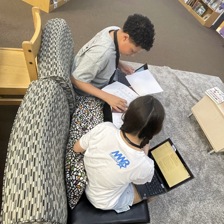 high-angle shot of two students working collaboratively at a library table. They are surrounded by printed research papers and are using laptops to type up their findings.