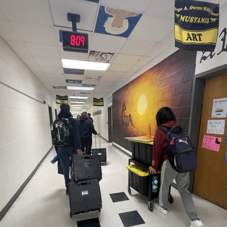 Students in a school hallway at Hugo A. Owens Middle School wheeling large black equipment bins and carts toward the exit. A digital clock overhead reads 8:09, and a sunset mural featuring a horse decorates the wall.