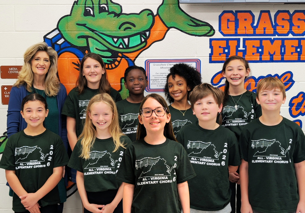A group of nine elementary school students and one adult stand smiling in front of a colorful school wall mural featuring a cartoon alligator and the words “Grassfield Elementary.” The students are wearing matching dark green T-shirts that read “All Virginia Elementary Chorus 2026.” They are arranged in two rows, with five students in front and four in the back alongside the adult. The group appears diverse, and everyone is facing the camera with cheerful expressions.