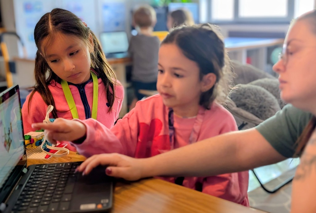 A picture of a teacher's arm and two student's wearing pink shirts.  One of the students is pointing to toward the Chromebook screen showing her teacher something while the other student looks on with concentration as they learn how  to avigate the software to control the LEGO robotic model sitting on the table between the two students.