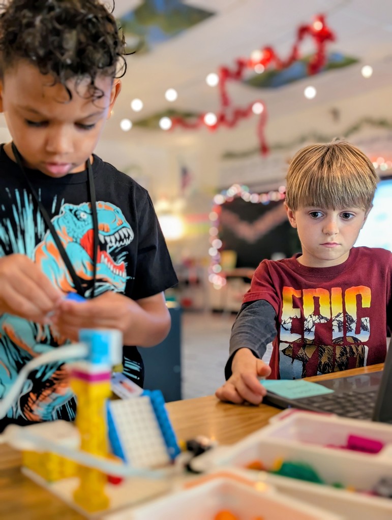 A close-up, of two boys working on a STEM project. The boy on the left, wearing a dinosaur T-shirt, adjusts a LEGO structure with wires. The boy on the right, in a maroon "Epic" shirt, watches the laptop screen with a serious, focused expression. String lights glow in the blurred background.