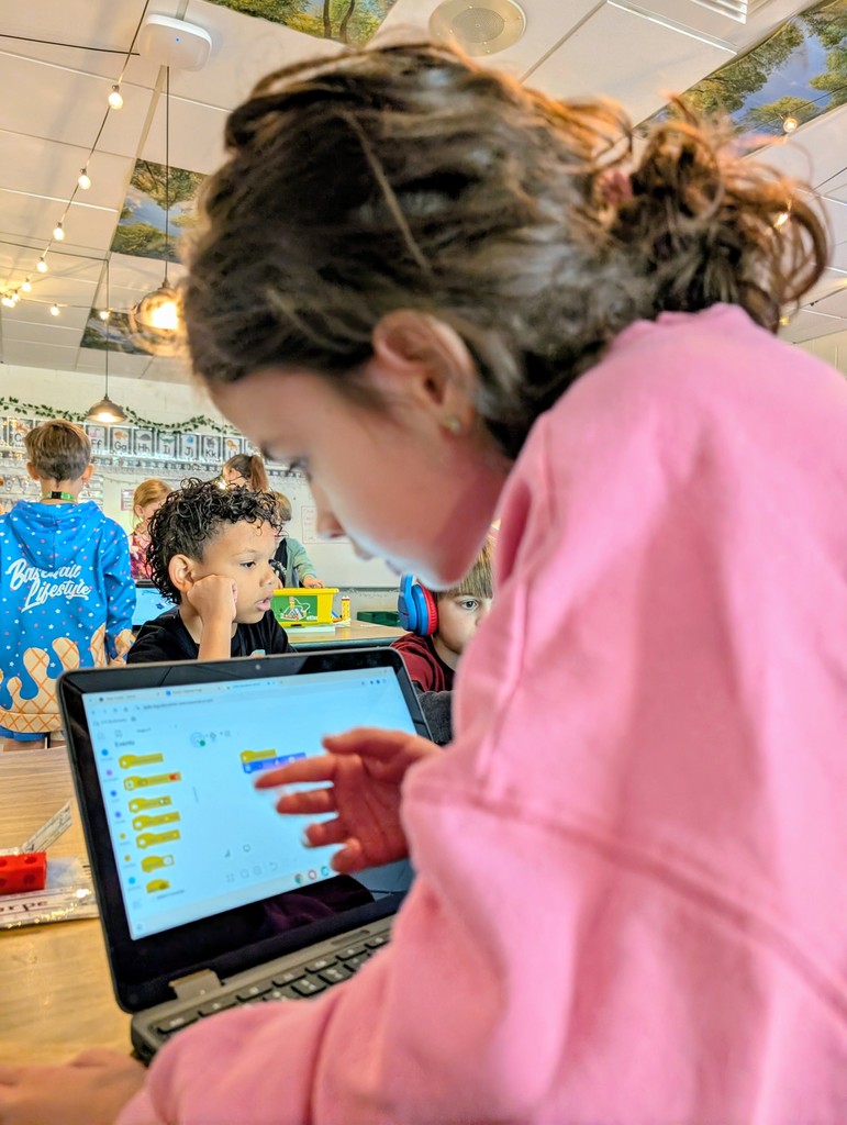 An over-the-shoulder shot of a young girl in a pink sweatshirt pointing at a laptop screen displaying a block-based coding interface. In the blurred background, another young boy rests his chin on his hand while looking toward the same workstation in a brightly lit classroom with string lights.