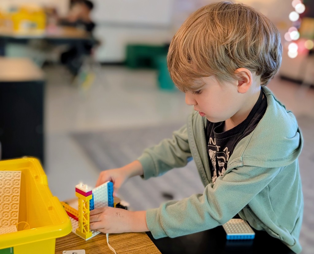 A young boy in a sage green zip-up hoodie works independently, carefully attaching a white and blue LEGO plate to a yellow support structure on a wooden table. A yellow bin of sorted LEGO parts is visible to his left.