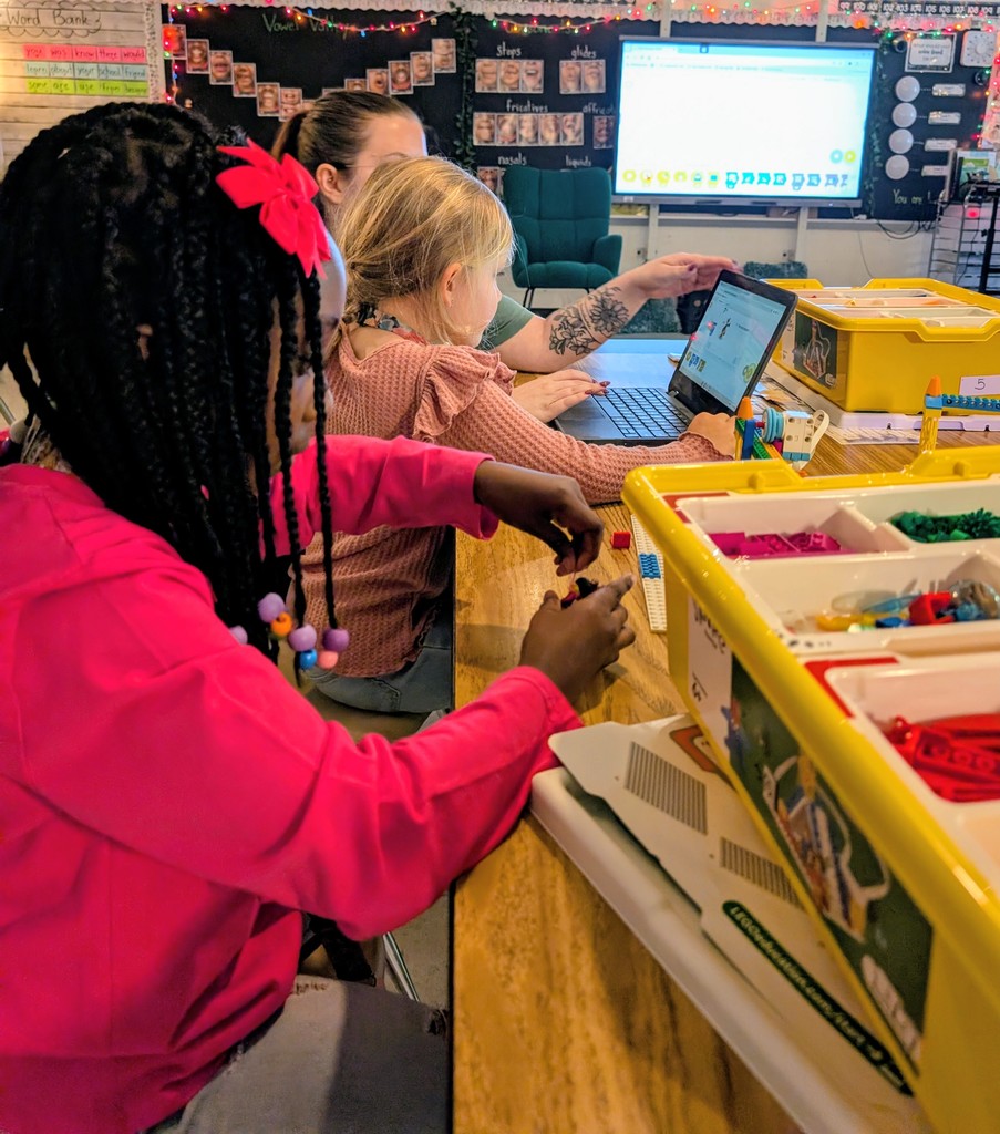 A side-view of a diverse classroom where a young girl in a bright pink hoodie works with LEGO Education bricks in the foreground. In the background, another student and a teacher look at a laptop screen together. Yellow storage bins filled with colorful bricks sit on the  table.