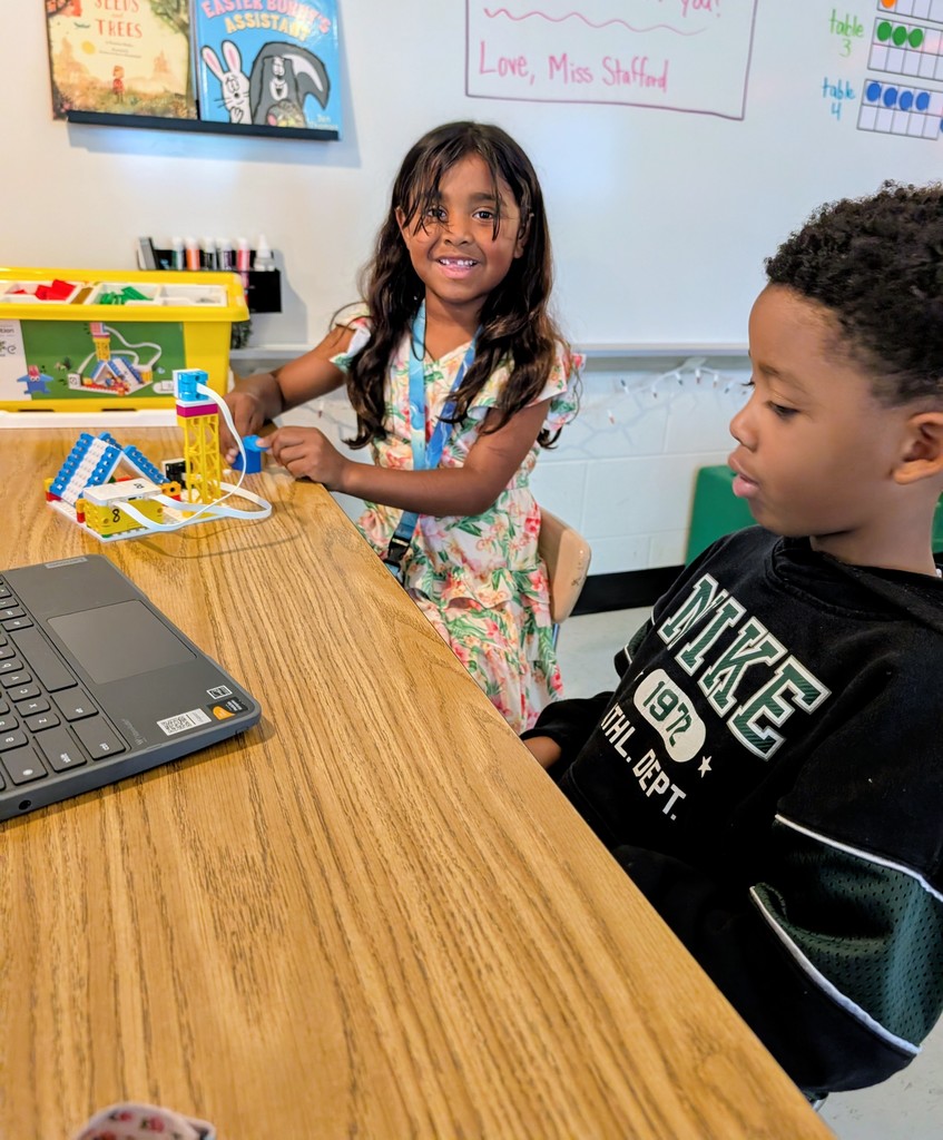 Two children sitting at a table; a girl in a floral dress smiles at the camera while holding a blue LEGO piece, and a boy in a black Nike hoodie looks intently at a Chromebook. Between them is a constructed LEGO model .