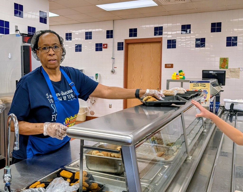 A medium shot of school nutrition professionals serving students behind a stainless steel counter.