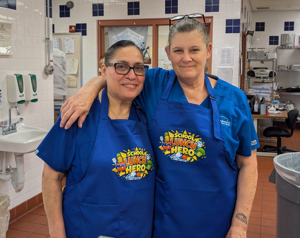 A joyful picture of two of GFE's cafeteria workers.  They are both wearing royal blue aprons with an image saying School Lunch Hero in bright bold orange and yellow colors.