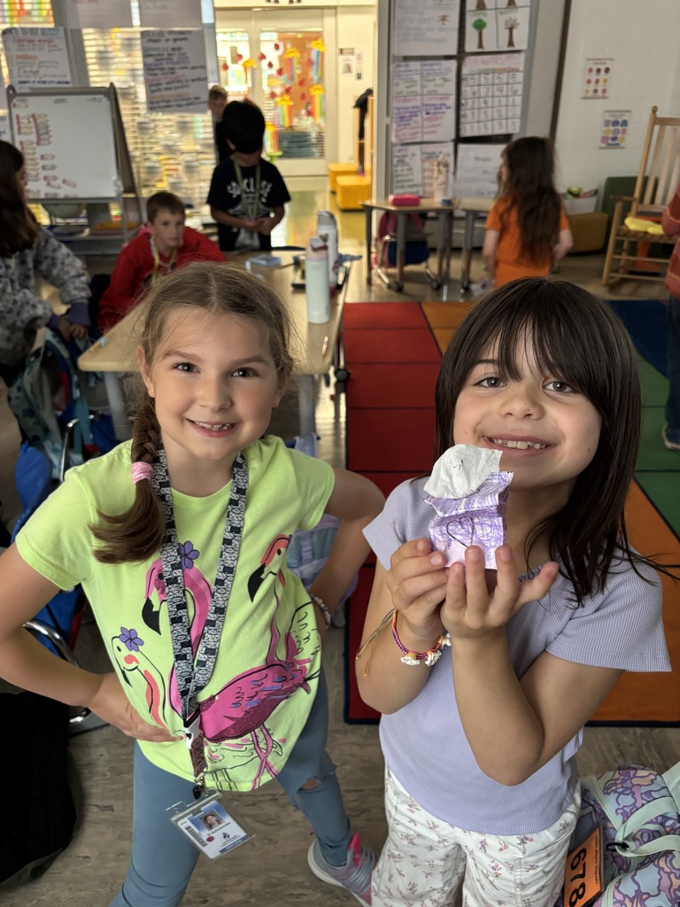 Two students smiling and holding up a real-world example of a solid figure, demonstrating their understanding of 3D shapes during a hands-on math activity in the classroom.