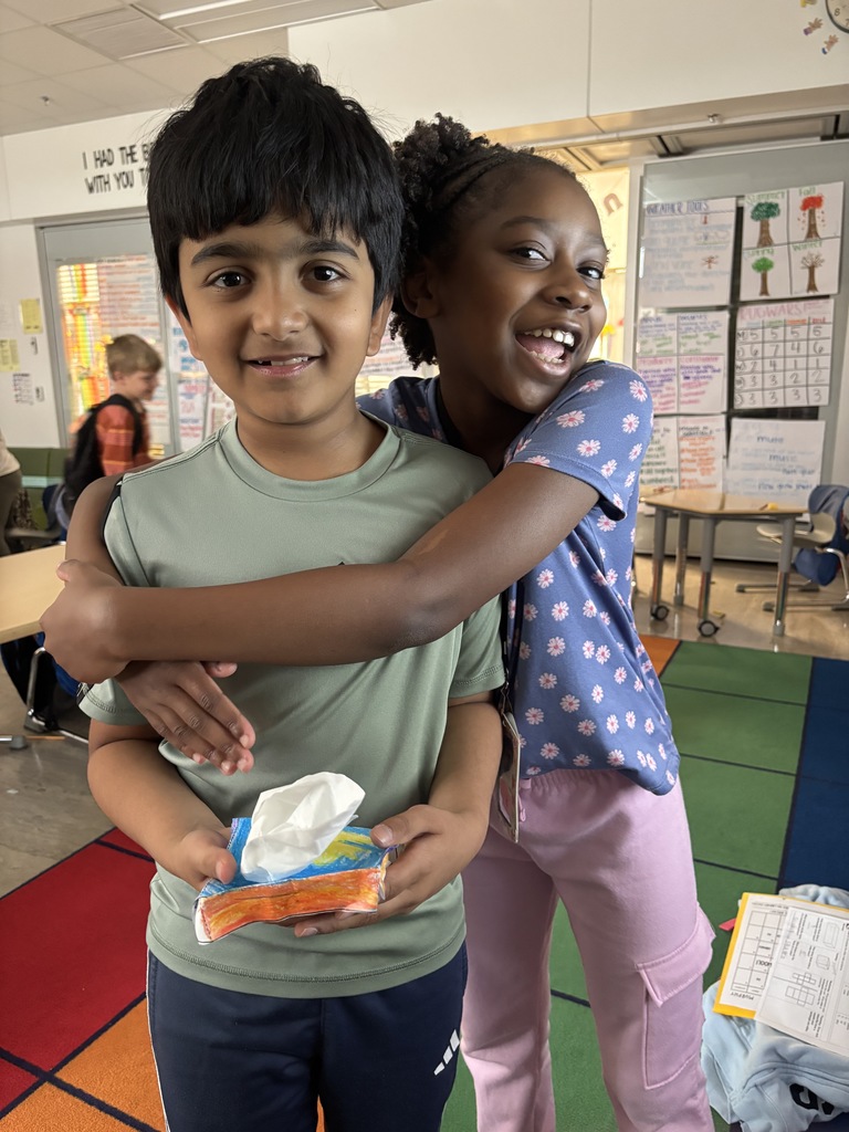 Two students smiling and holding up a real-world example of a solid figure, demonstrating their understanding of 3D shapes during a hands-on math activity in the classroom.