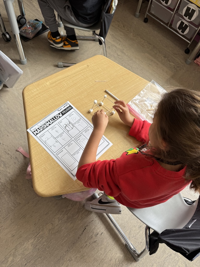 A student carefully building a cube model using toothpicks and marshmallows, connecting the pieces to form a 3D shape during a hands-on math activity.