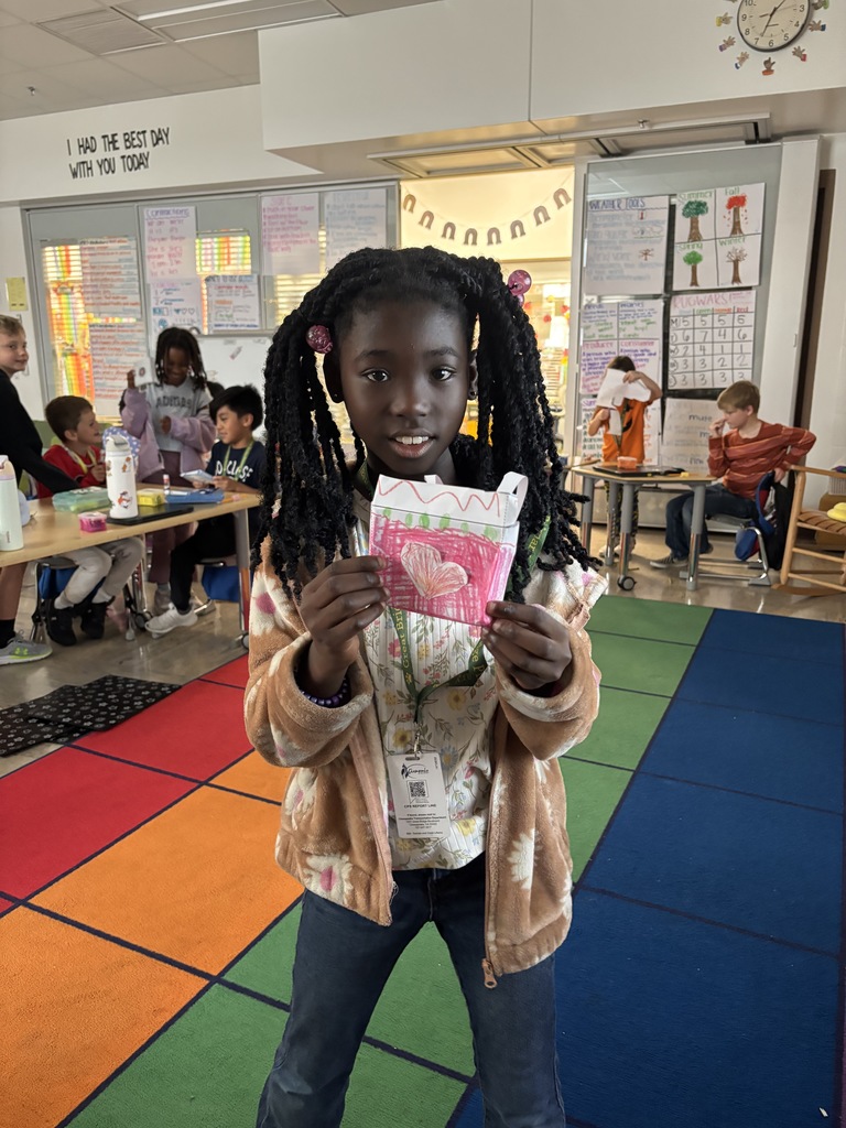 A student smiling and holding up a real-world example of a solid figure, demonstrating their understanding of 3D shapes during a hands-on math activity in the classroom.