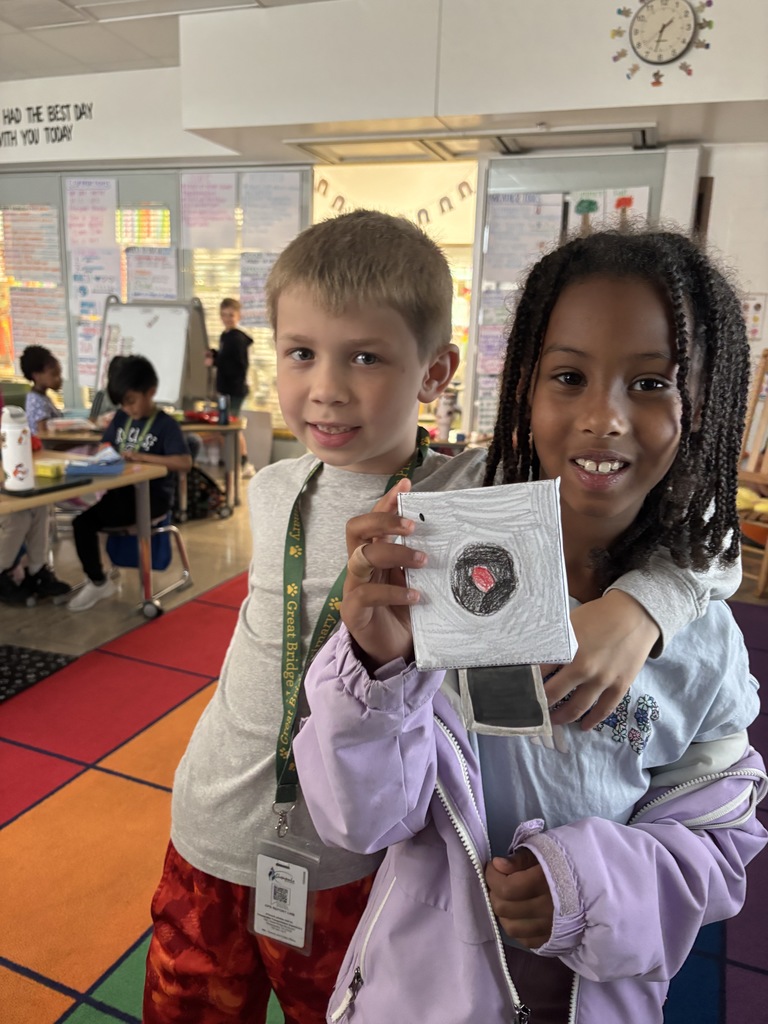 Two students smiling and holding up a real-world example of a solid figure, demonstrating their understanding of 3D shapes during a hands-on math activity in the classroom.