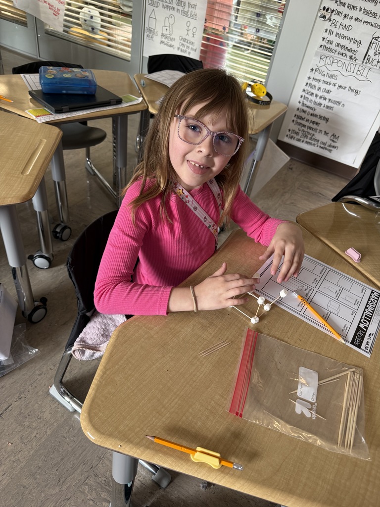A student smiling as she is carefully building a cube model using toothpicks and marshmallows, connecting the pieces to form a 3D shape during a hands-on math activity.